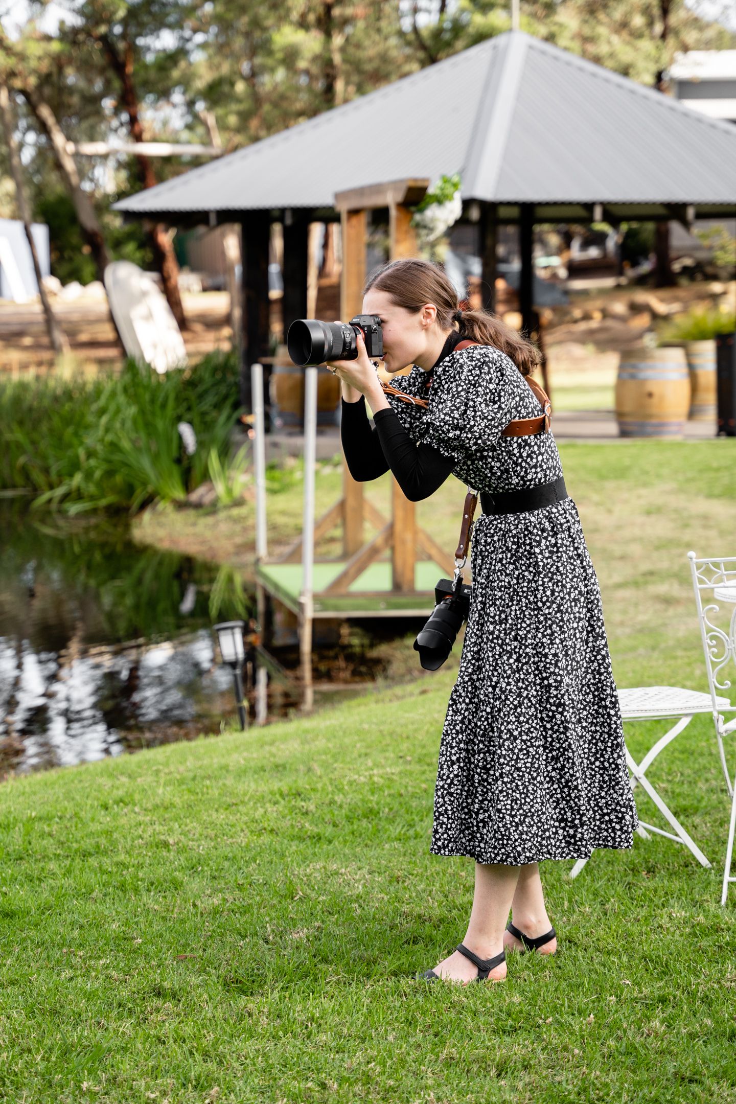 Event photographer in a patterned dress taking photos by a pond with a gazebo in the background.
