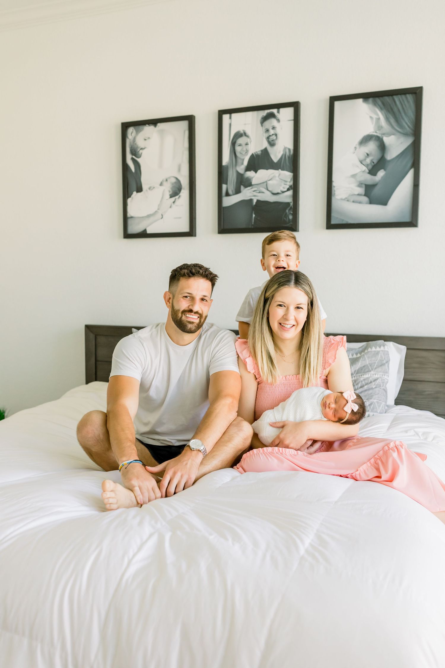 A happy family poses together on their bed with black and white photos displayed behind them.