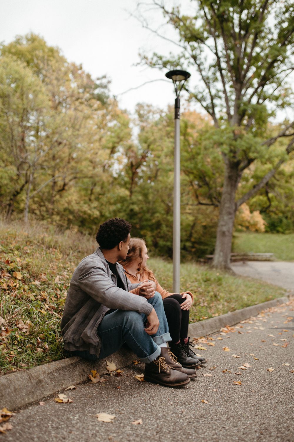 A couple sits together on a curb beneath a lamppost in a park setting.