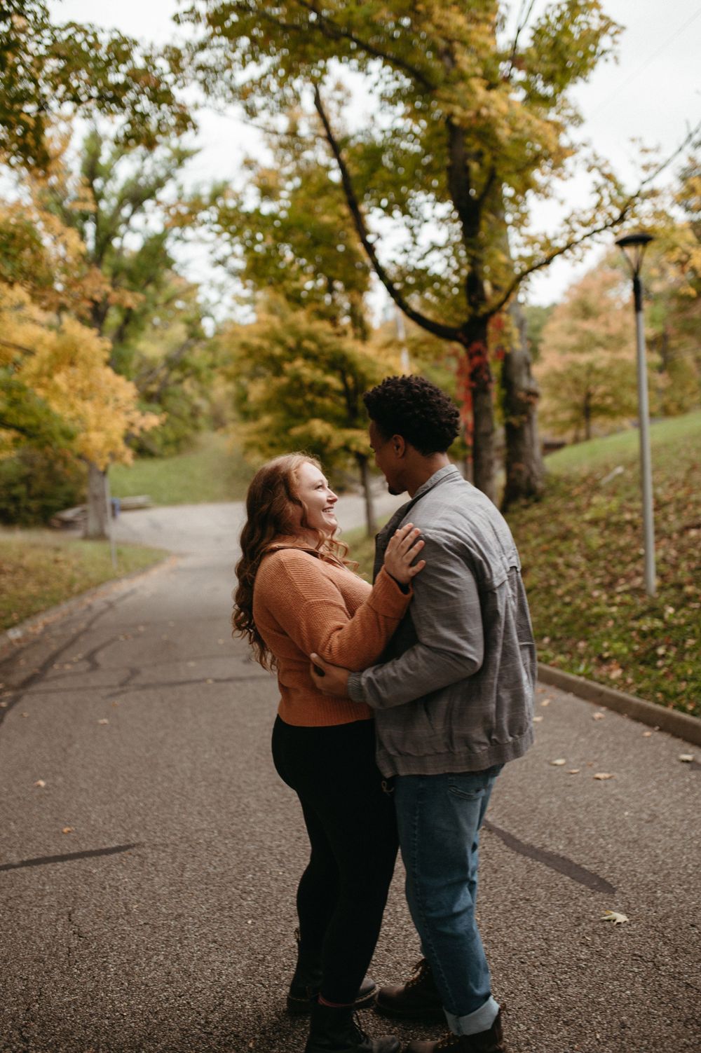 A couple shares an intimate moment on a park path surrounded by autumn trees.