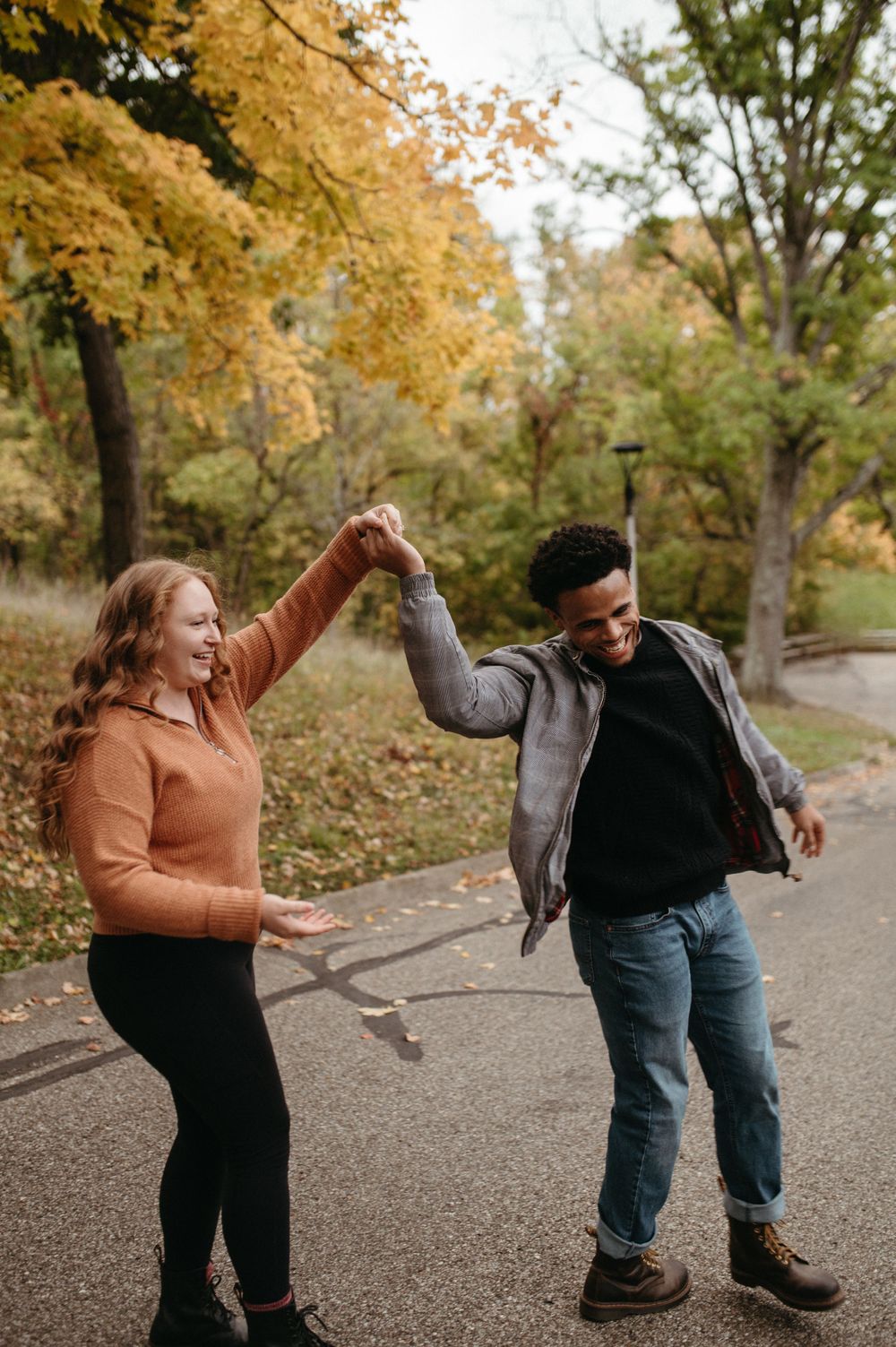 A couple holds hands and dances joyfully on a park path during fall.