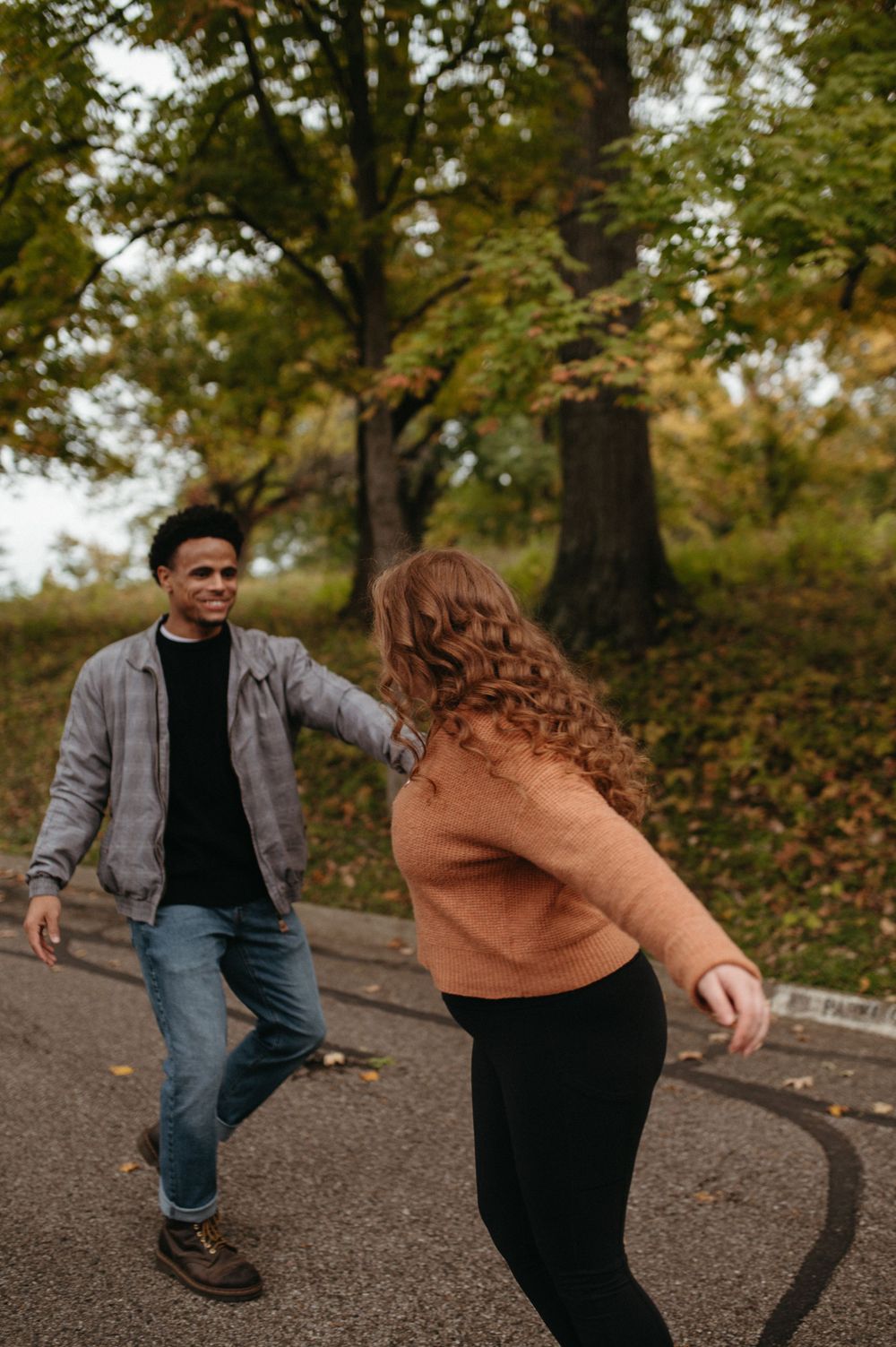 A couple dances playfully on a paved pathway surrounded by autumn foliage.