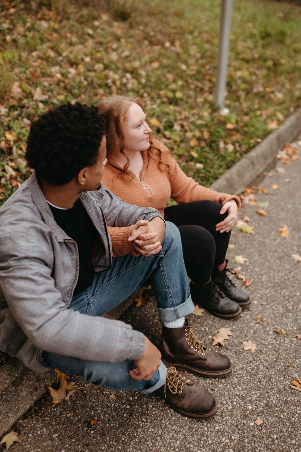 A couple sits close together on a curb surrounded by fallen autumn leaves.