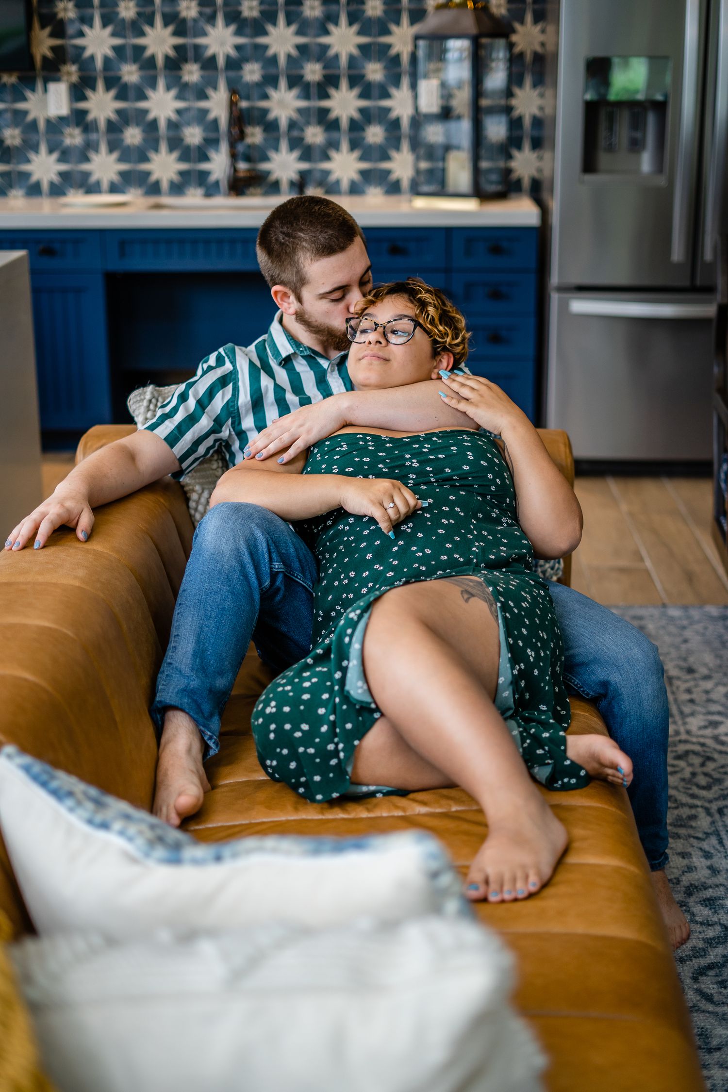 A couple relaxes on a tan leather couch in a modern kitchen with blue patterned tile.