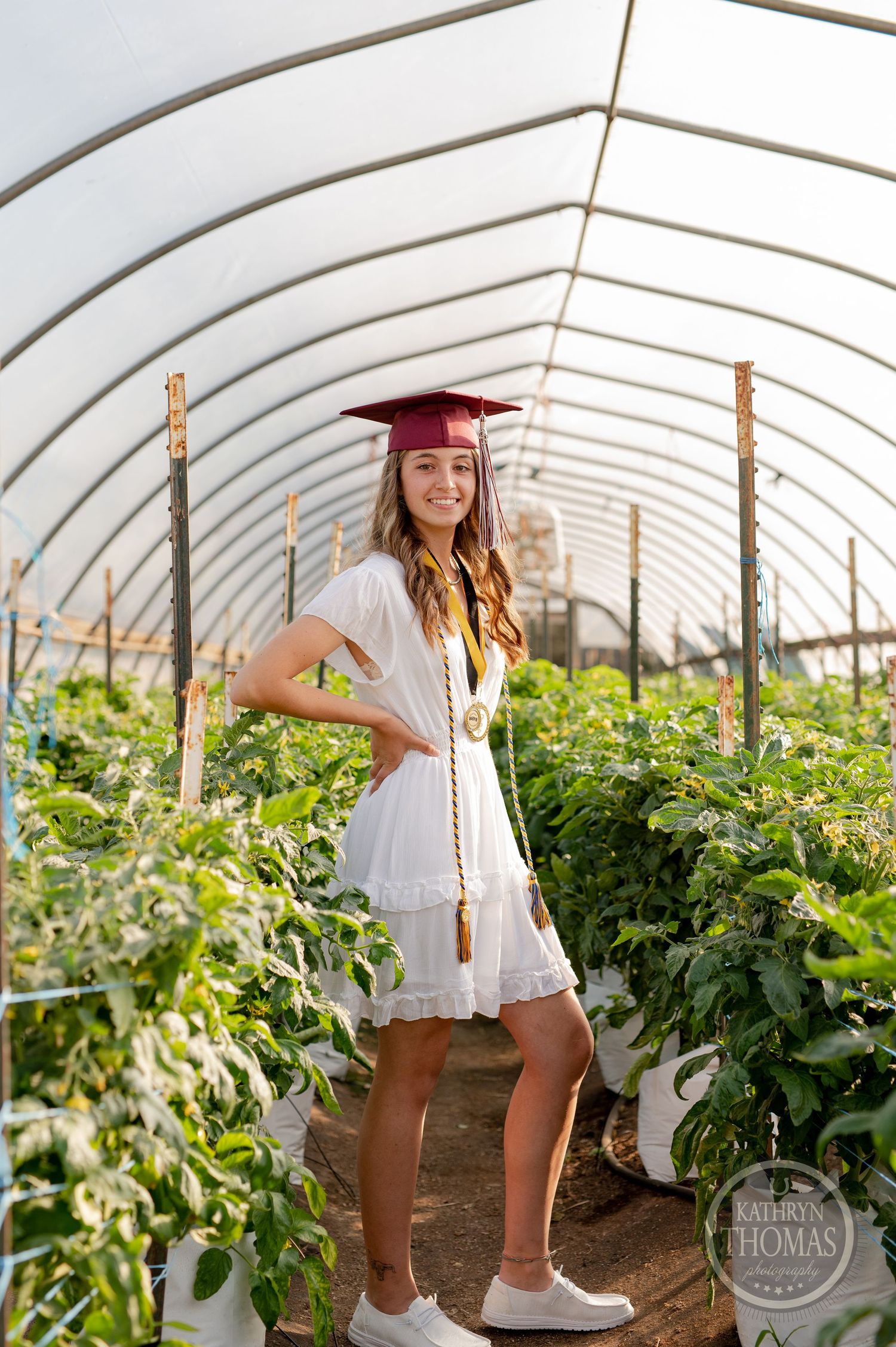 Graduate stands among lush greenhouse plants wearing white dress and maroon graduation cap.
