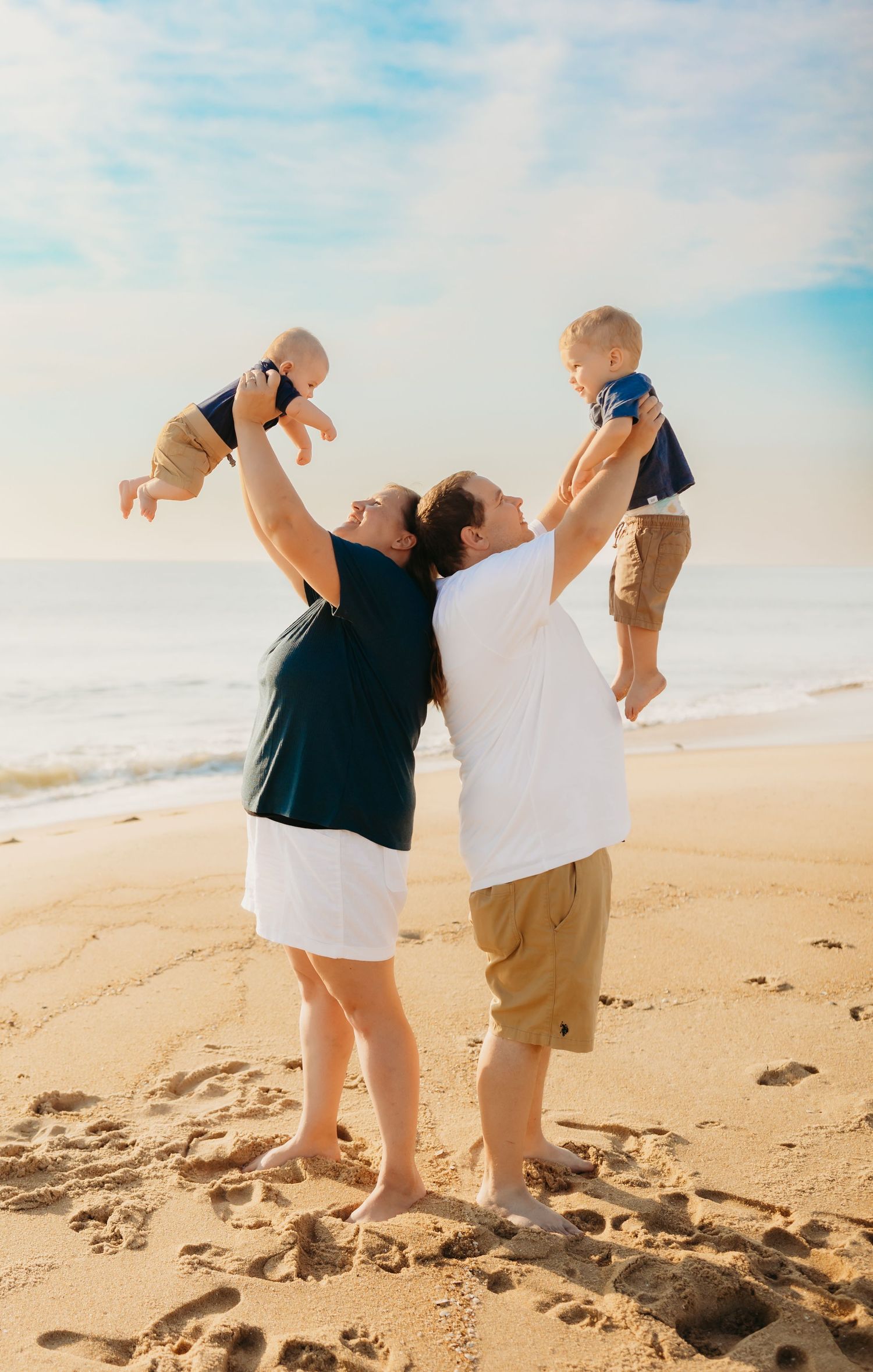 Joyful parents lifting two young children on a sunny beach with ocean waves in the background.