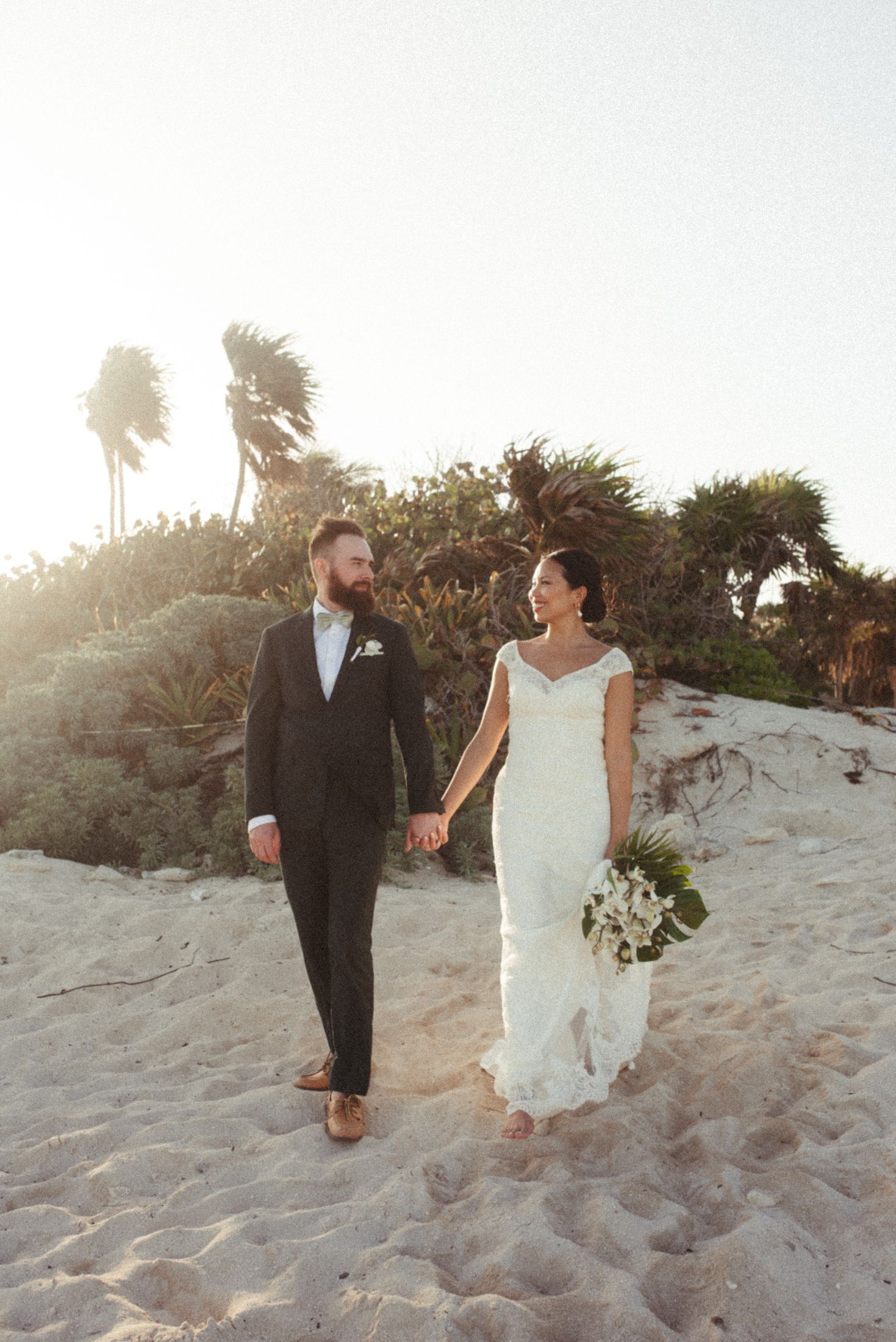 A couple walks hand in hand on a sandy beach at sunset, with palm trees in the background.