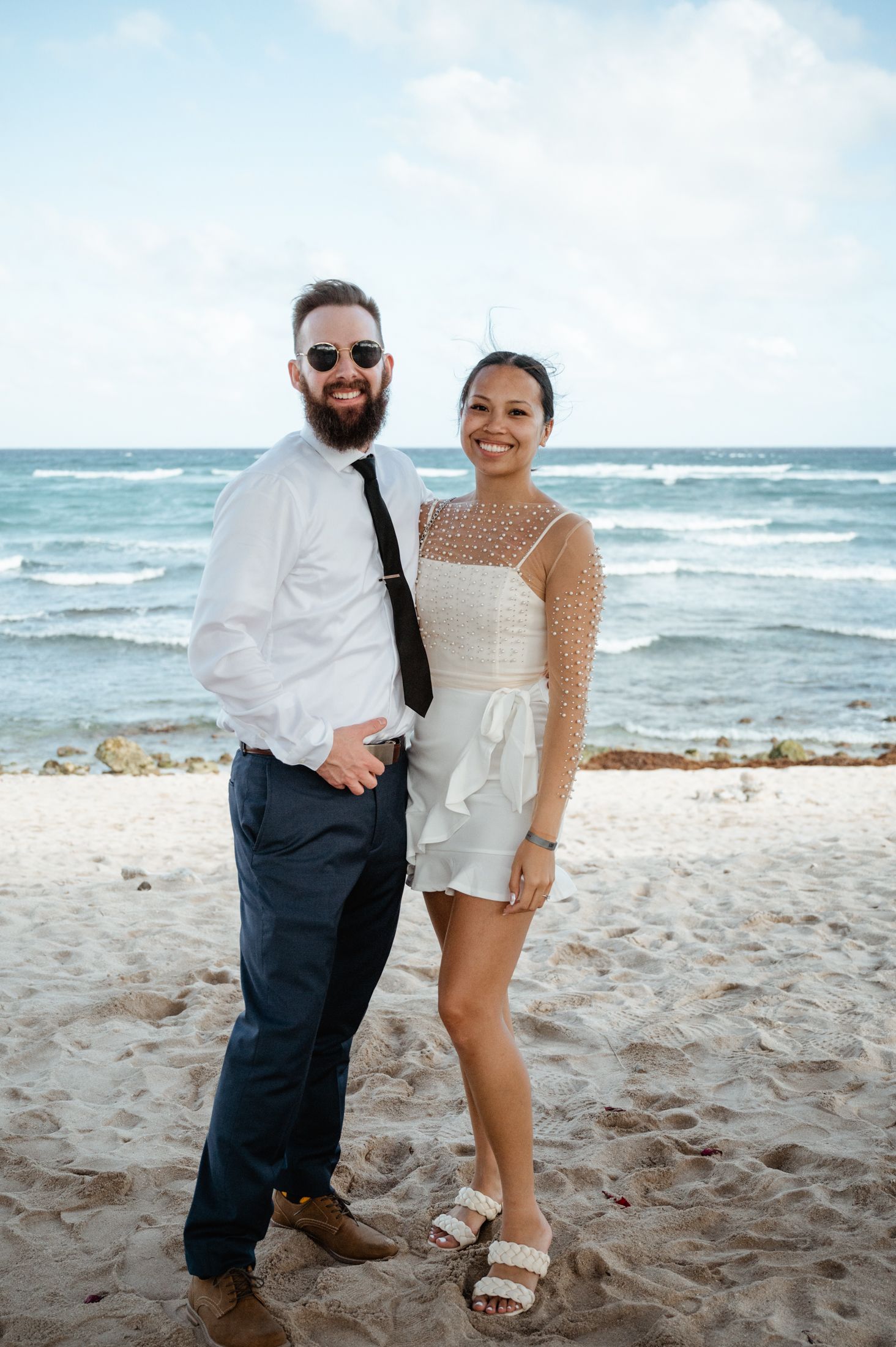 A couple poses together on a beach with ocean waves and cloudy sky in the background.