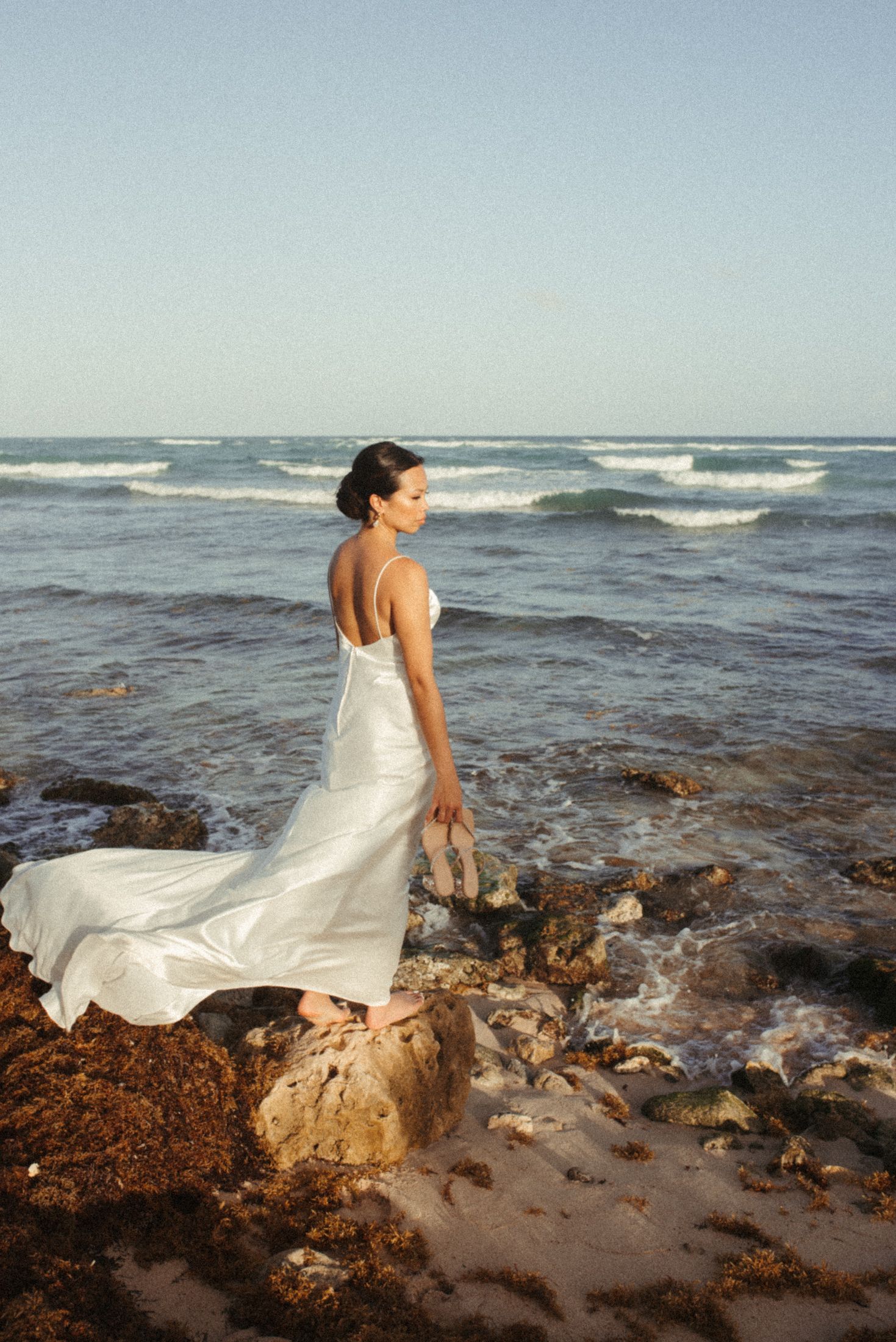 A bride in a white dress stands on rocky coastline facing the ocean waves.
