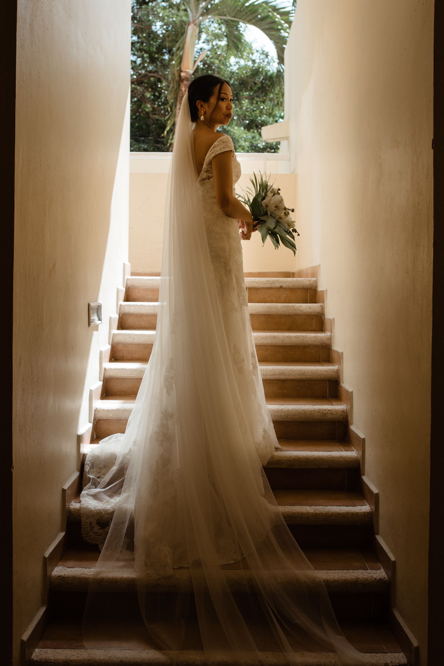 A bride in a flowing white gown poses on stone stairs in dramatic lighting.
