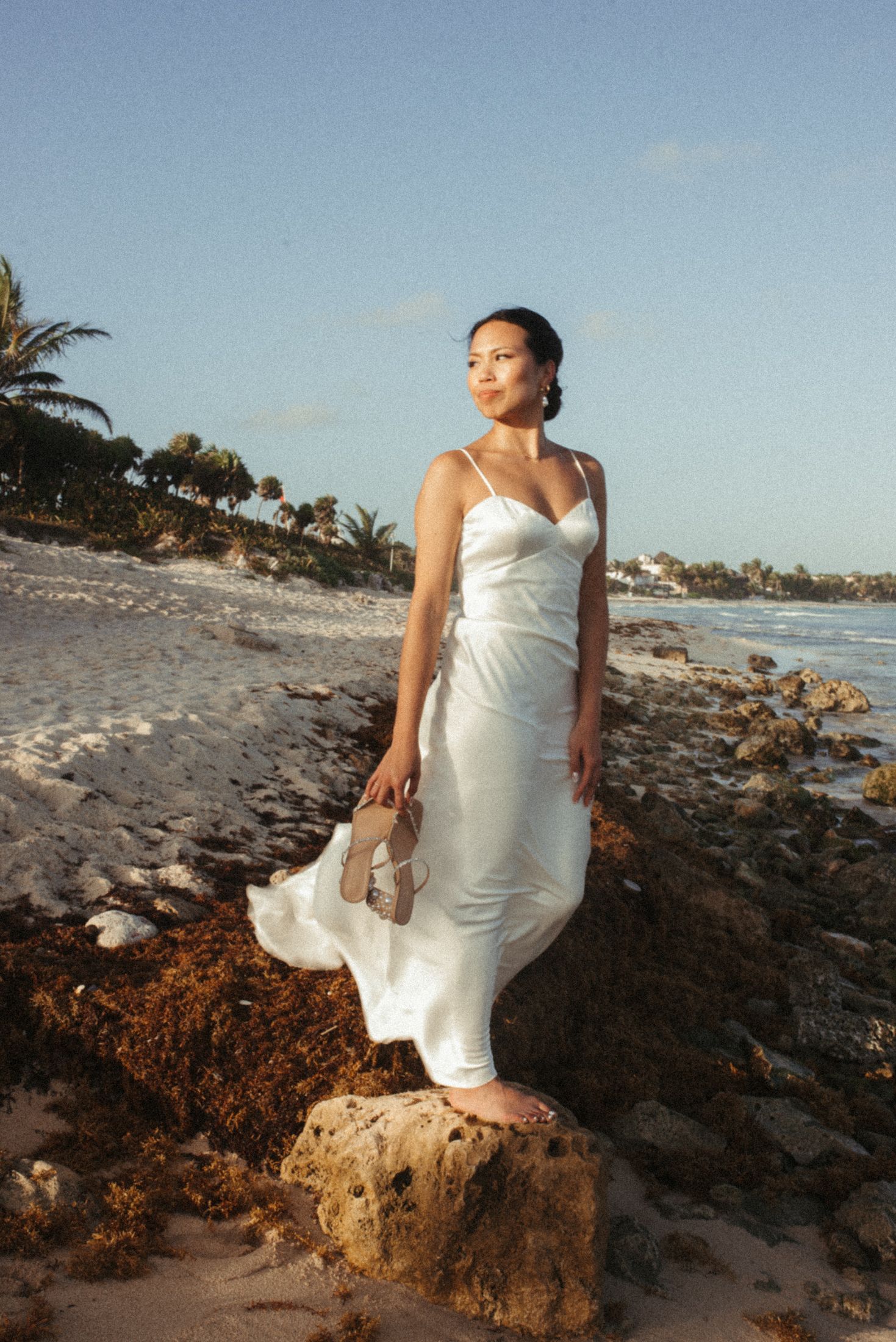 A bride in a flowing white dress poses on coastal rocks during golden hour.