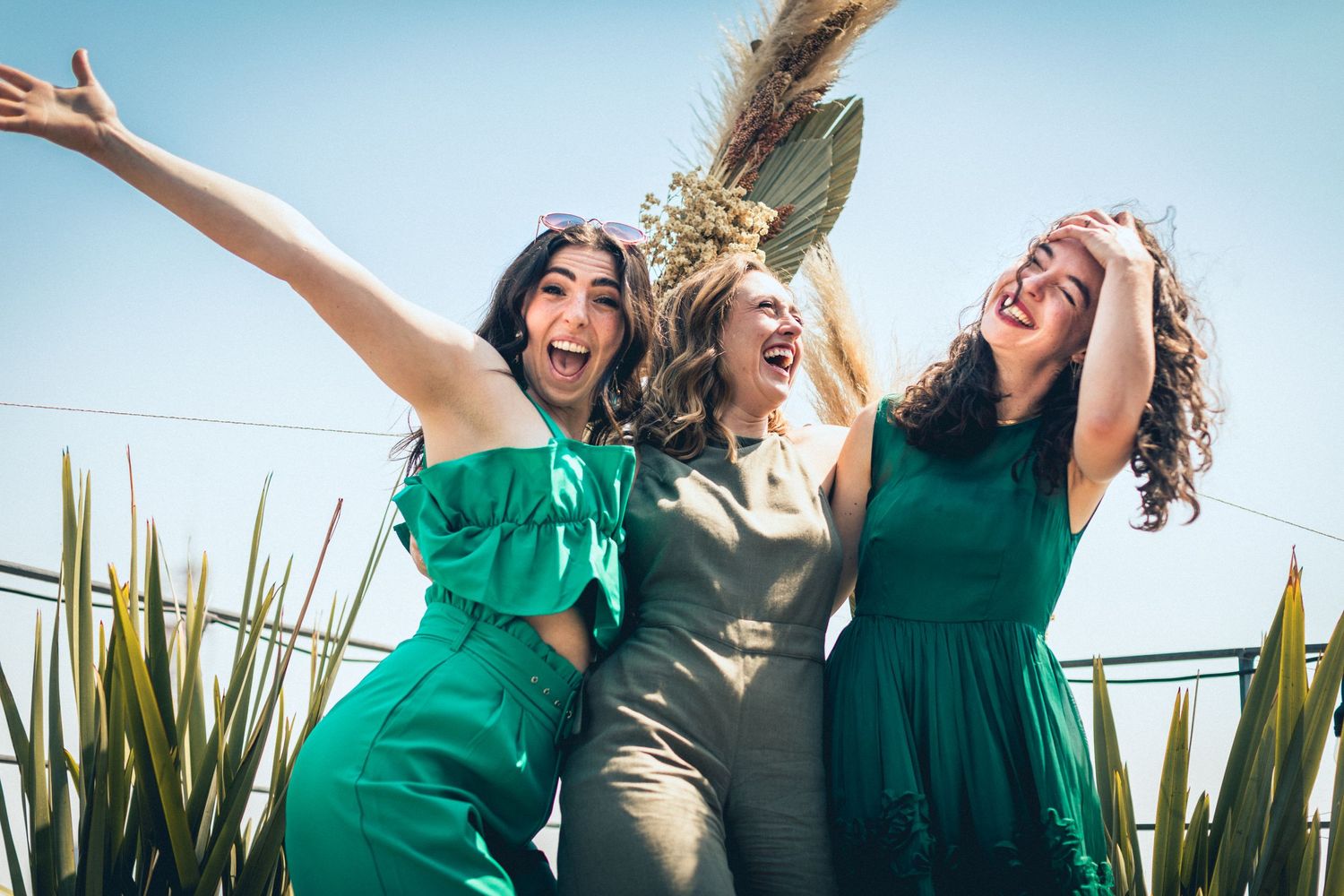 Wedding party poses energetically with bridesmaids in green dresses against blue sky backdrop.