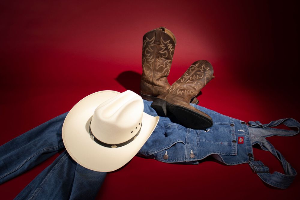 Western-style arrangement of cowboy boots, denim jeans, and white hat against red backdrop.