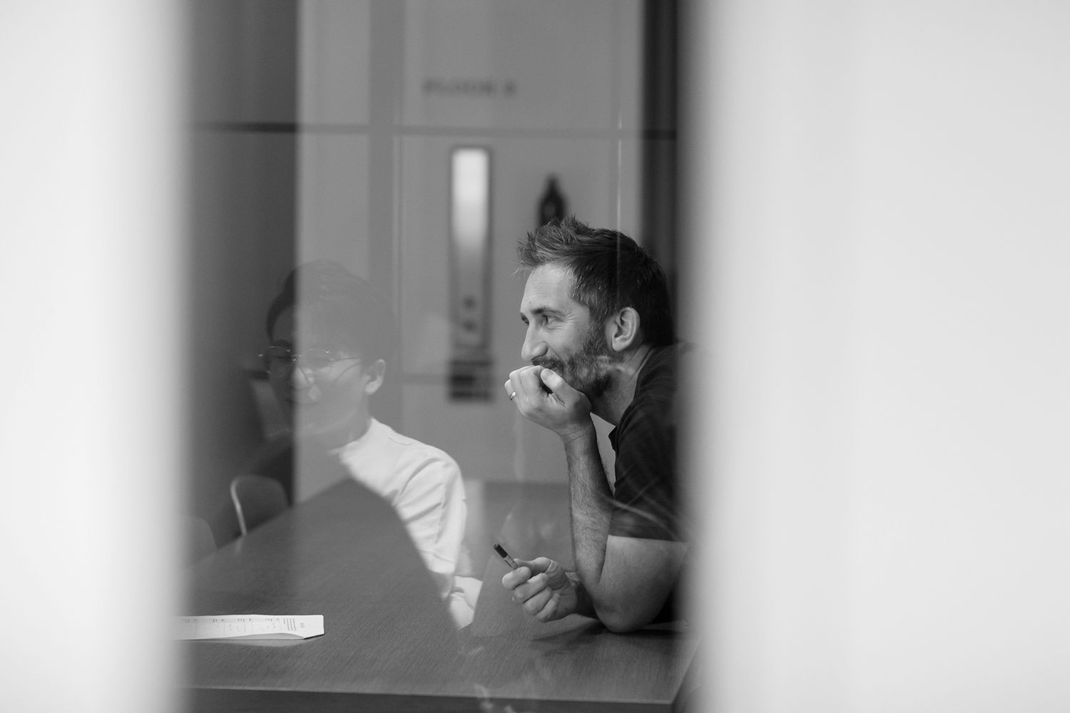 Black and white photo of a person in profile viewed through a window or glass partition during business branding photoshoot.