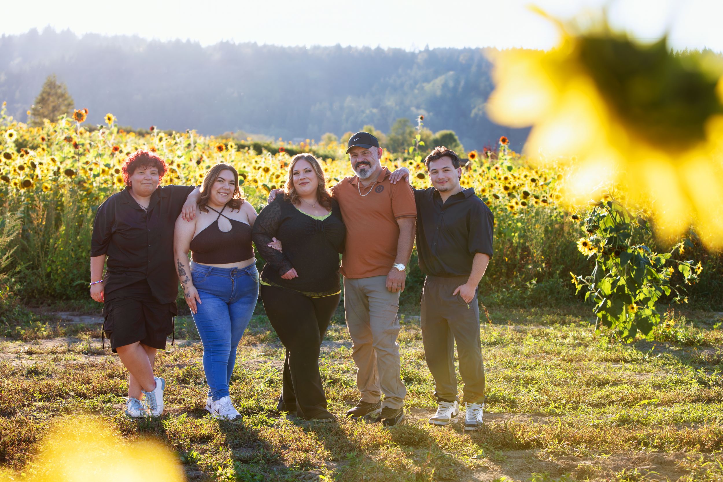 A group of friends stand together in a sunflower field at golden hour with mountains visible in the background.
