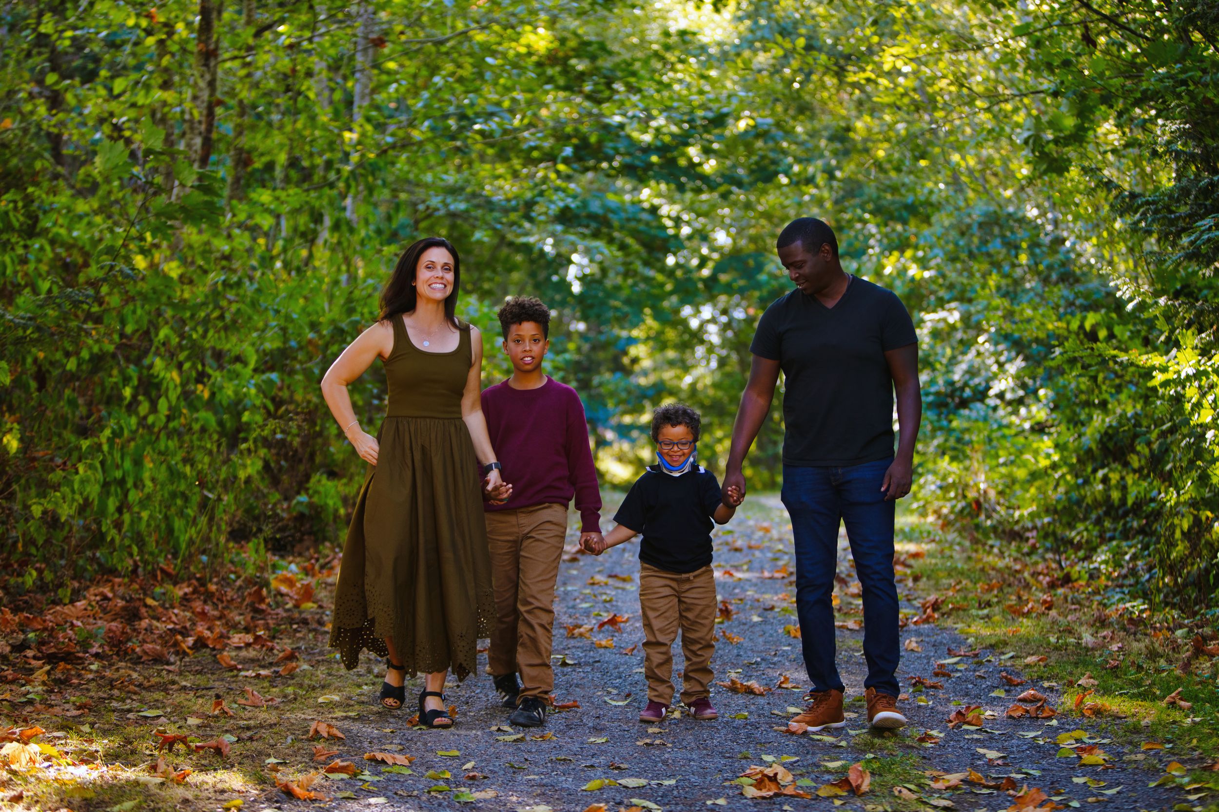 A group walks together holding hands along a scenic wooded path during autumn with fallen leaves on the ground.