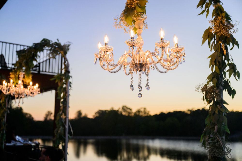Elegant crystal chandeliers hang from a rustic wooden pergola at an outdoor sunset wedding reception.