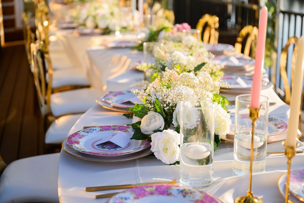 Elegant wedding table setting with pink taper candles, white floral centerpieces, and place settings on a long white tablecloth.