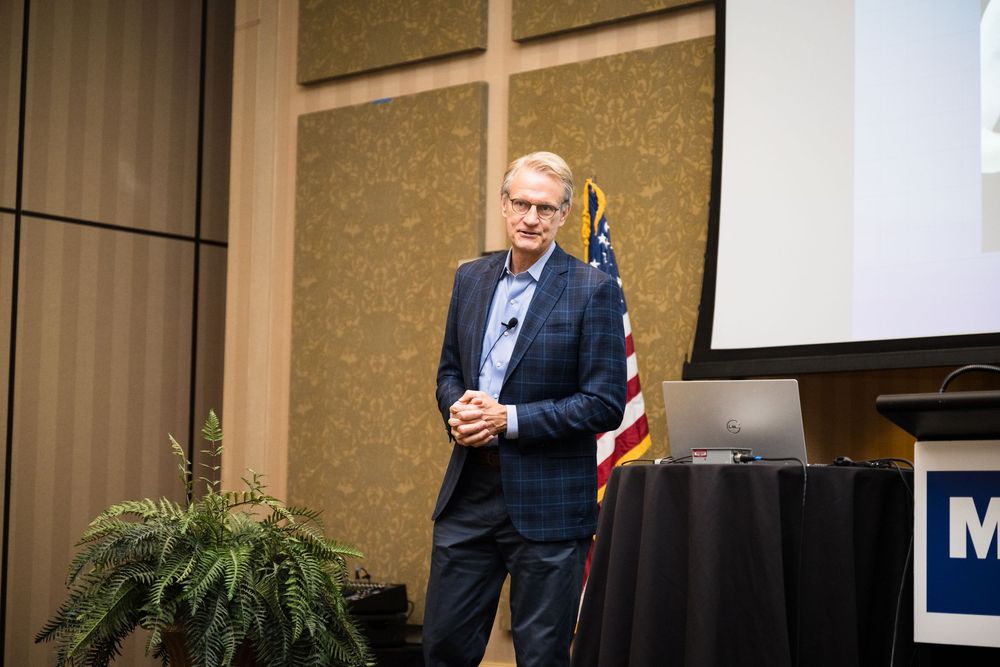 Professional speaker presenting at a business conference with projection screen and greenery on stage.