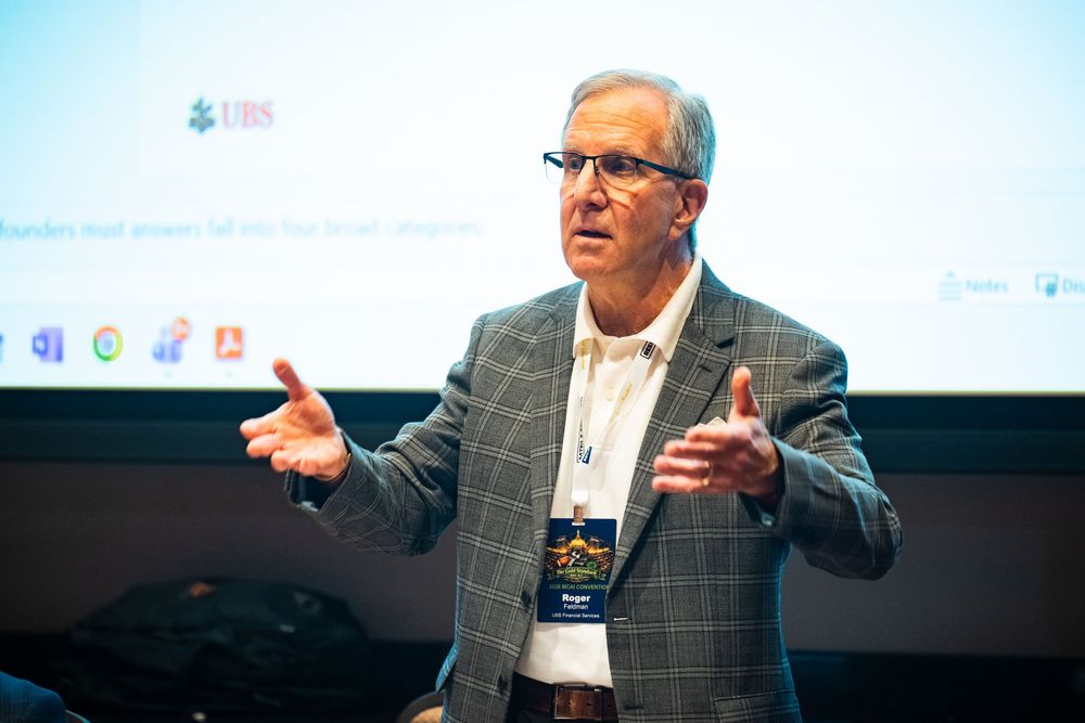 A man in a plaid blazer speaks passionately at a business conference, gesturing with both hands near a presentation screen.