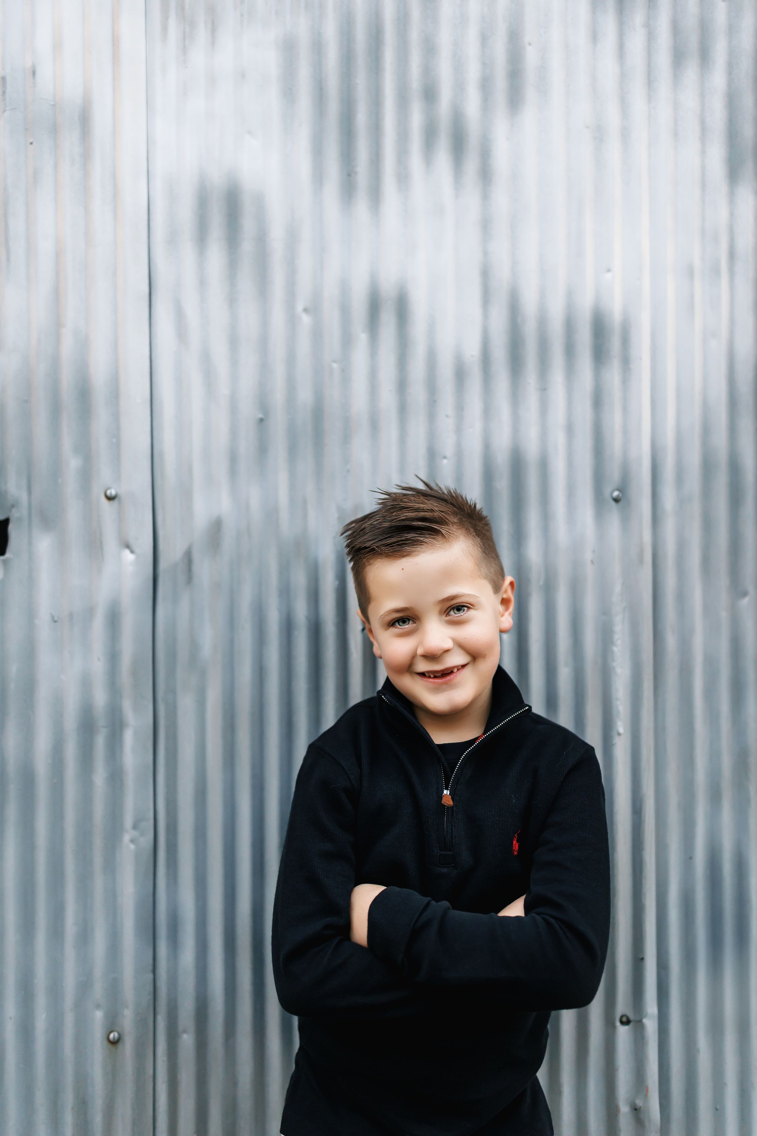 A child in a black hoodie poses playfully against a corrugated metal wall background.