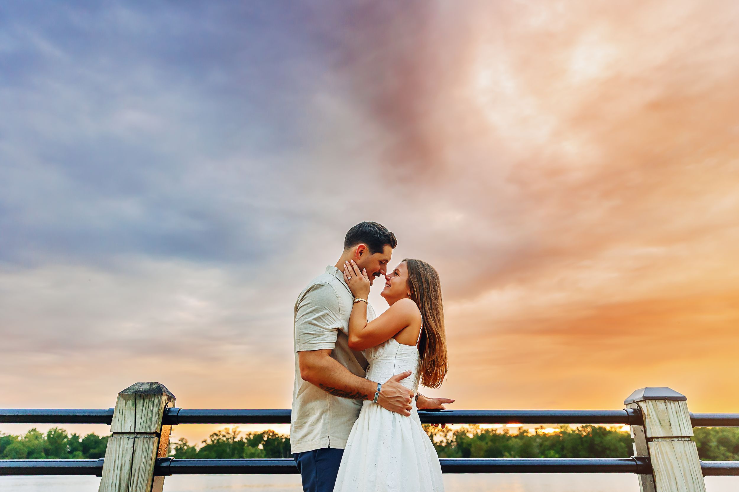 A couple shares a romantic kiss on a wooden pier at sunset with dramatic orange and pink clouds in the sky.