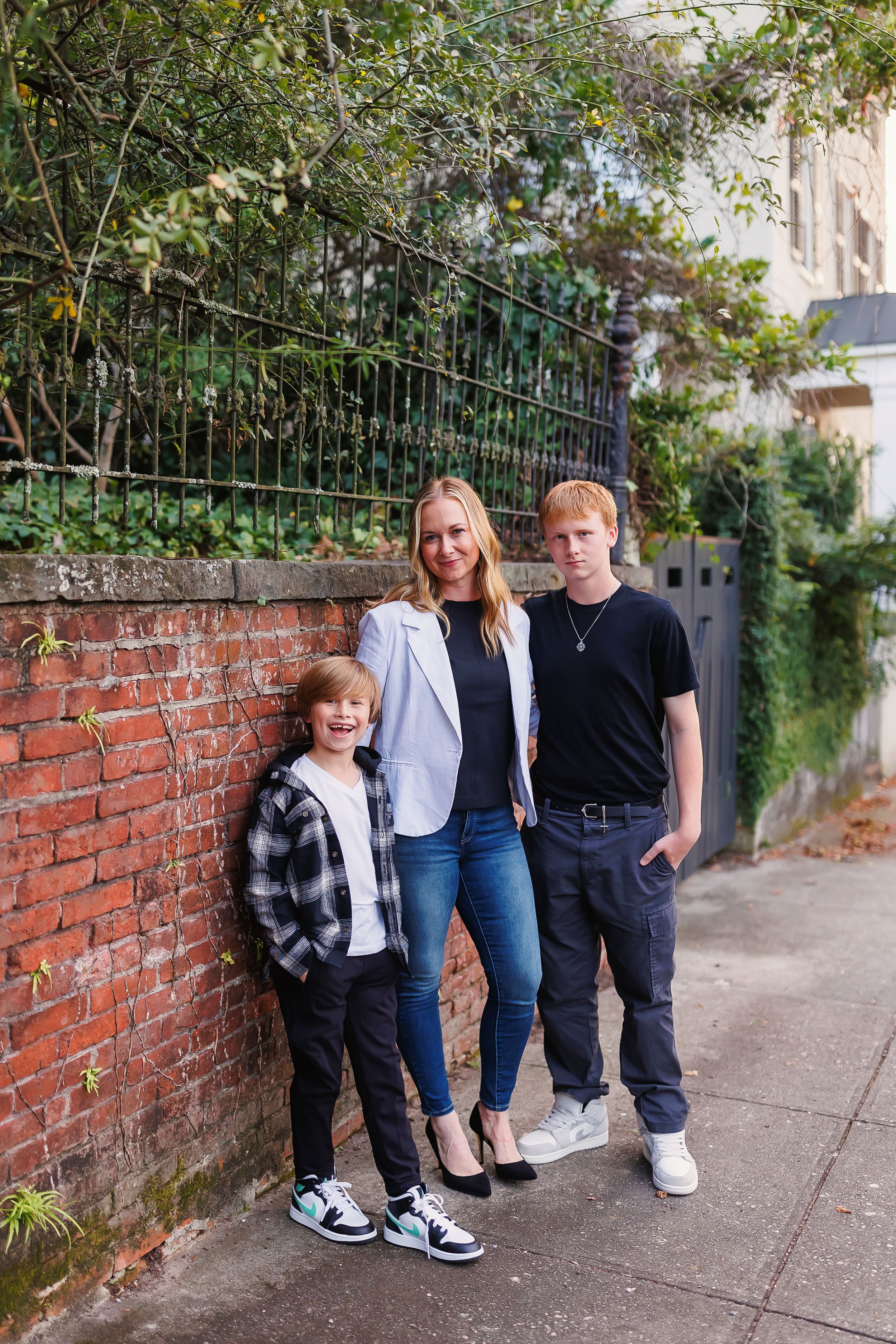 A group stands casually against a brick wall with wrought iron fence in an outdoor urban setting.