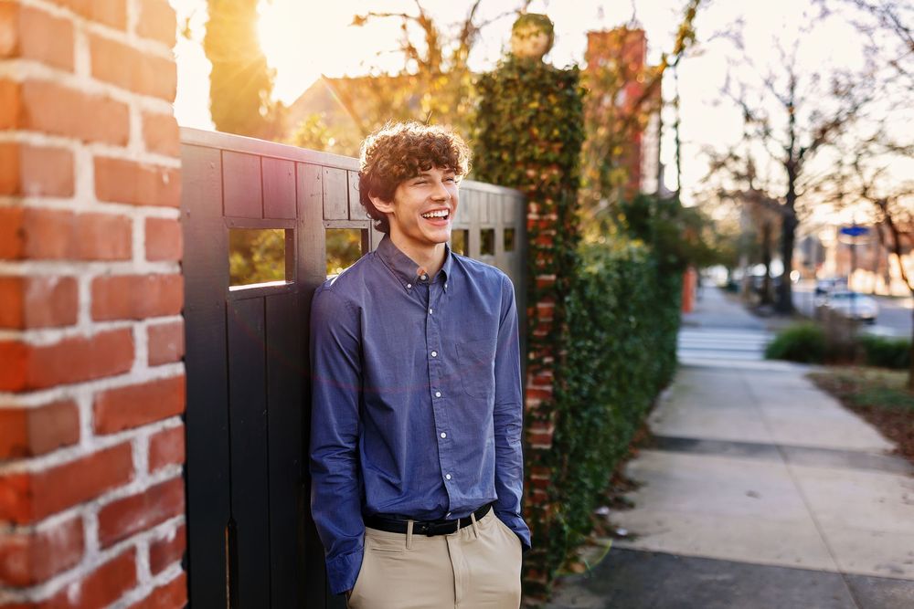 Young person in blue button-down shirt and khakis leaning against brick wall at sunset on city sidewalk.
