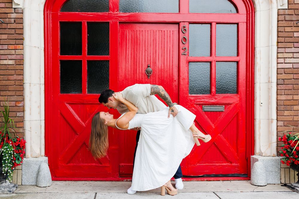 A romantic couple in white attire dips in front of a vibrant red door with brick walls and potted flowers.