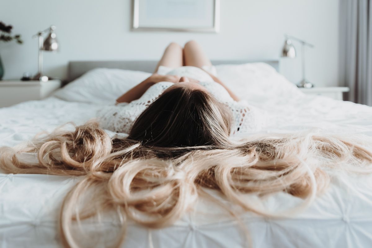 A person lying on a white bed with long blonde hair spread across the pillow.