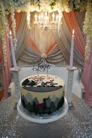 Elegant wedding cake with painted mountain scene and Love topper flanked by candles under chandelier.