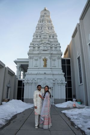 Couple in traditional Indian wedding attire posing before ornate white Hindu temple with gold statue.