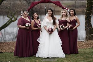 Bride in white gown with four bridesmaids in burgundy dresses holding bouquets by lakeside arch.