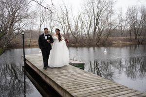 Bride and groom walking hand in hand on wooden dock beside calm lake with bare winter trees.