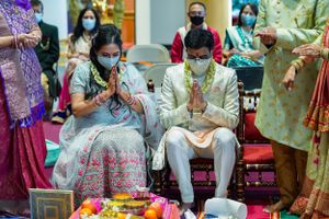 Couple in traditional Indian wedding attire performing prayer ceremony with guests wearing masks in background.