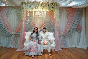 Couple in traditional South Asian attire seated on white sofa before elegant pink and mint floral draped backdrop.