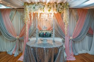 Elegant wedding cake display with pink and gray draped backdrop, floral arch, chandelier, and silver sequin tablecloth.