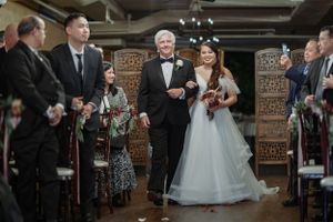 Bride in white gown walks down aisle with older gentleman at elegant indoor wedding ceremony with guests watching.