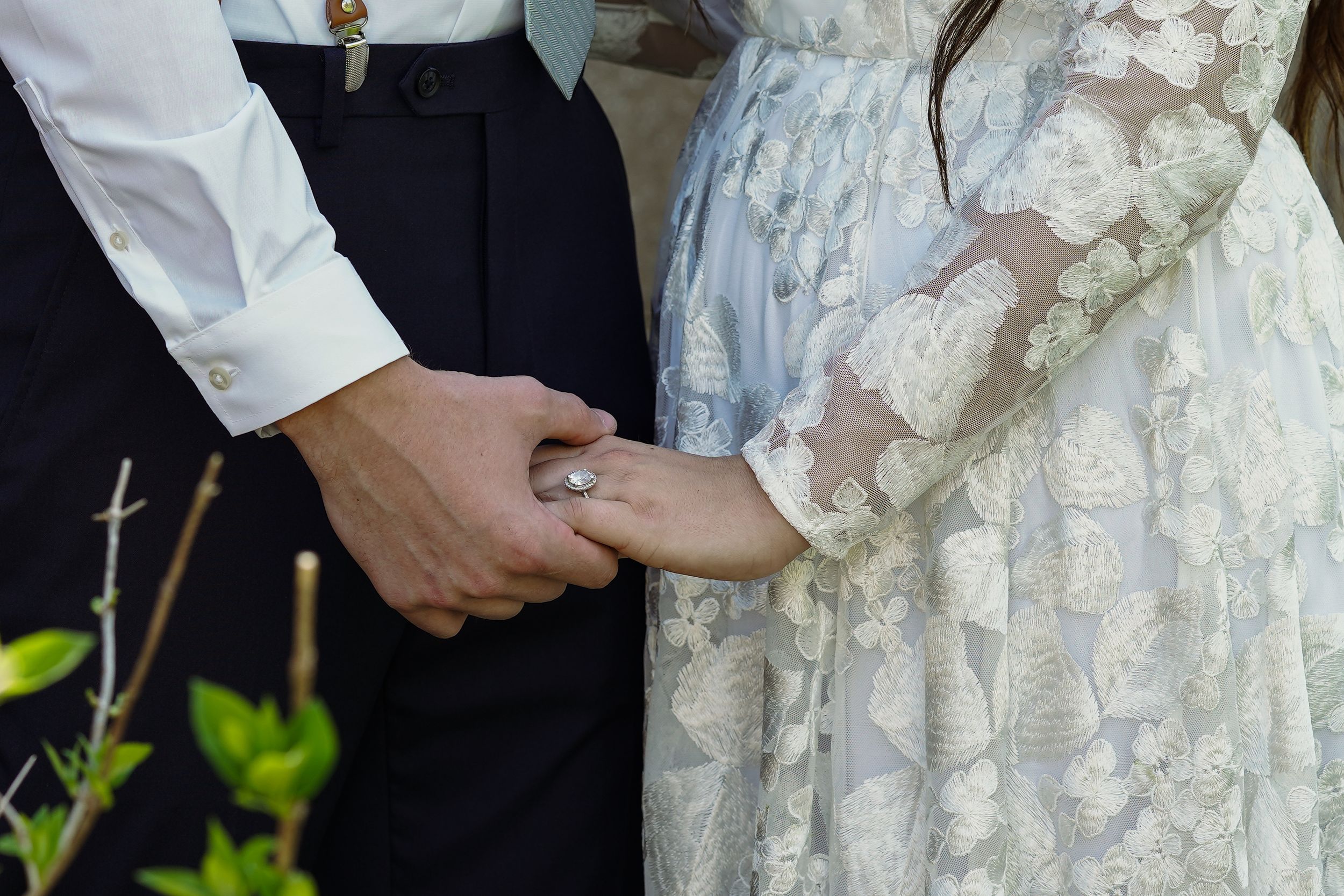 Couple holding hands at wedding ceremony, bride wearing lace floral dress with diamond ring, groom in white shirt and black pants.