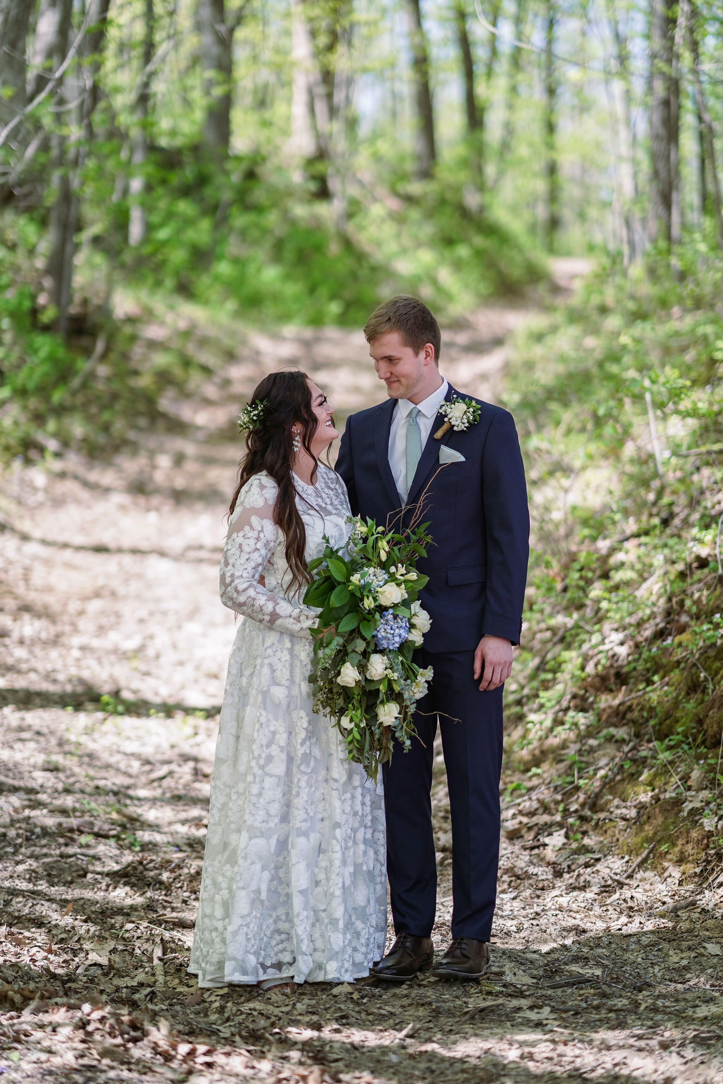 Bride and groom stand on a wooded forest path, she holds a lush bouquet, both in wedding attire gazing at each other.