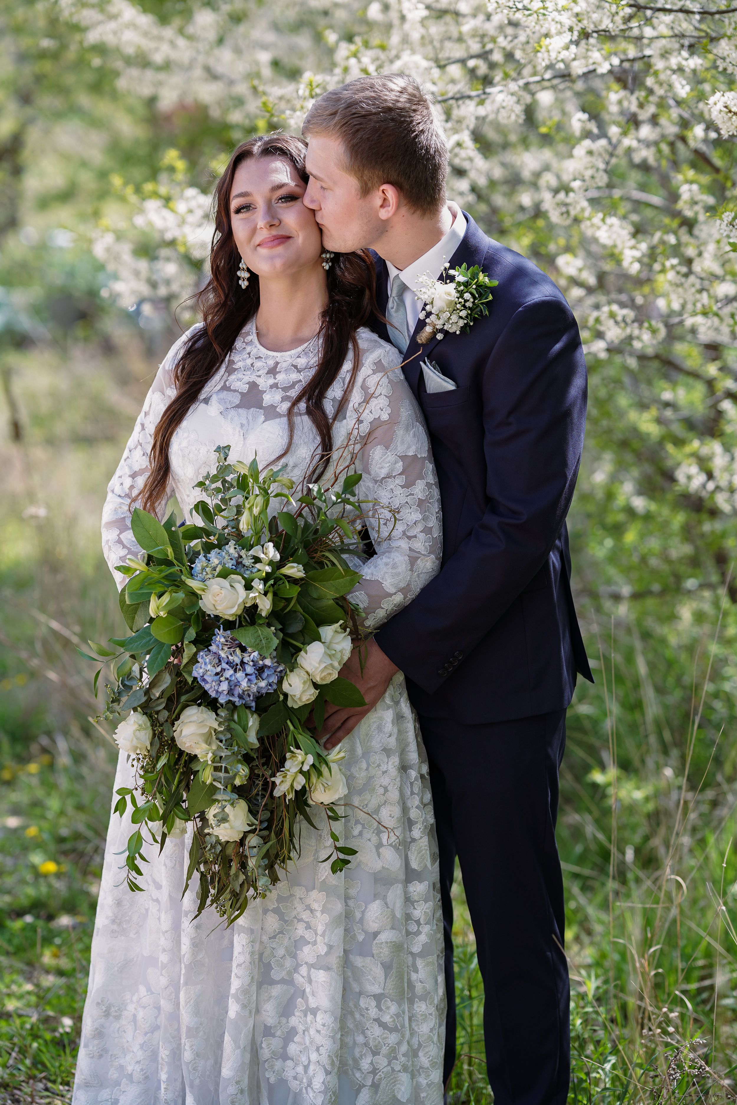 Groom in navy suit kisses bride in lace dress holding blue and white bouquet among blooming trees.