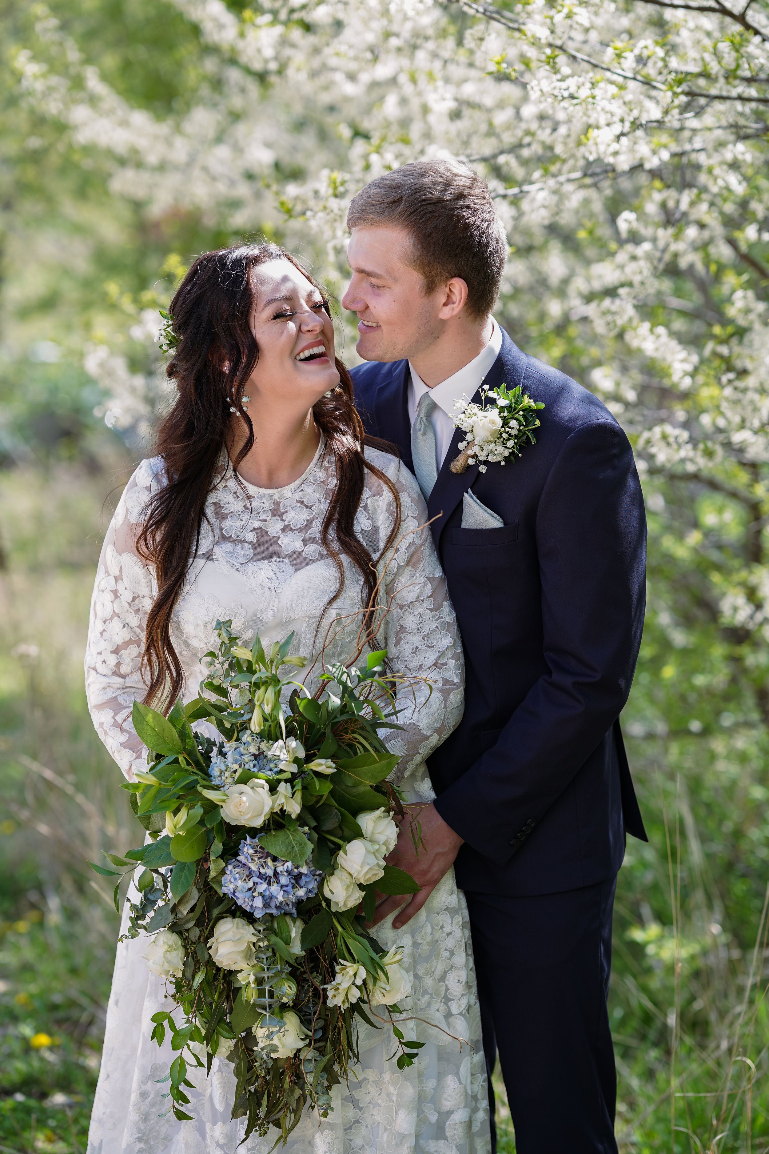 A smiling bride in lace dress holds blue floral bouquet beside groom in navy suit under blooming white cherry trees.