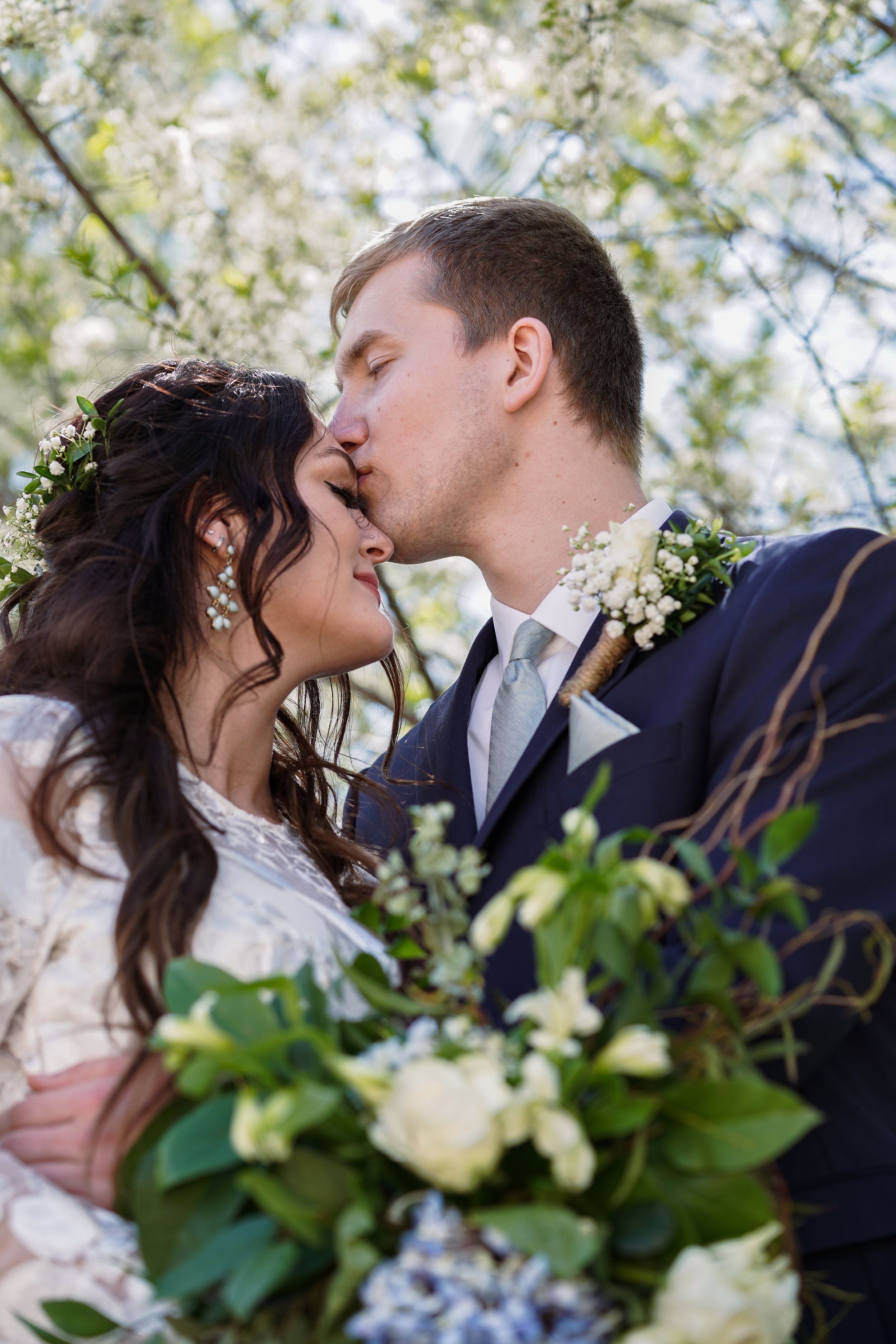 A bride and groom share a romantic kiss outdoors surrounded by white spring blossoms and a lush floral bouquet.