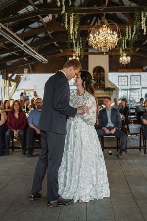 Bride and groom share first dance in rustic venue with chandeliers and seated guests watching during wedding reception.