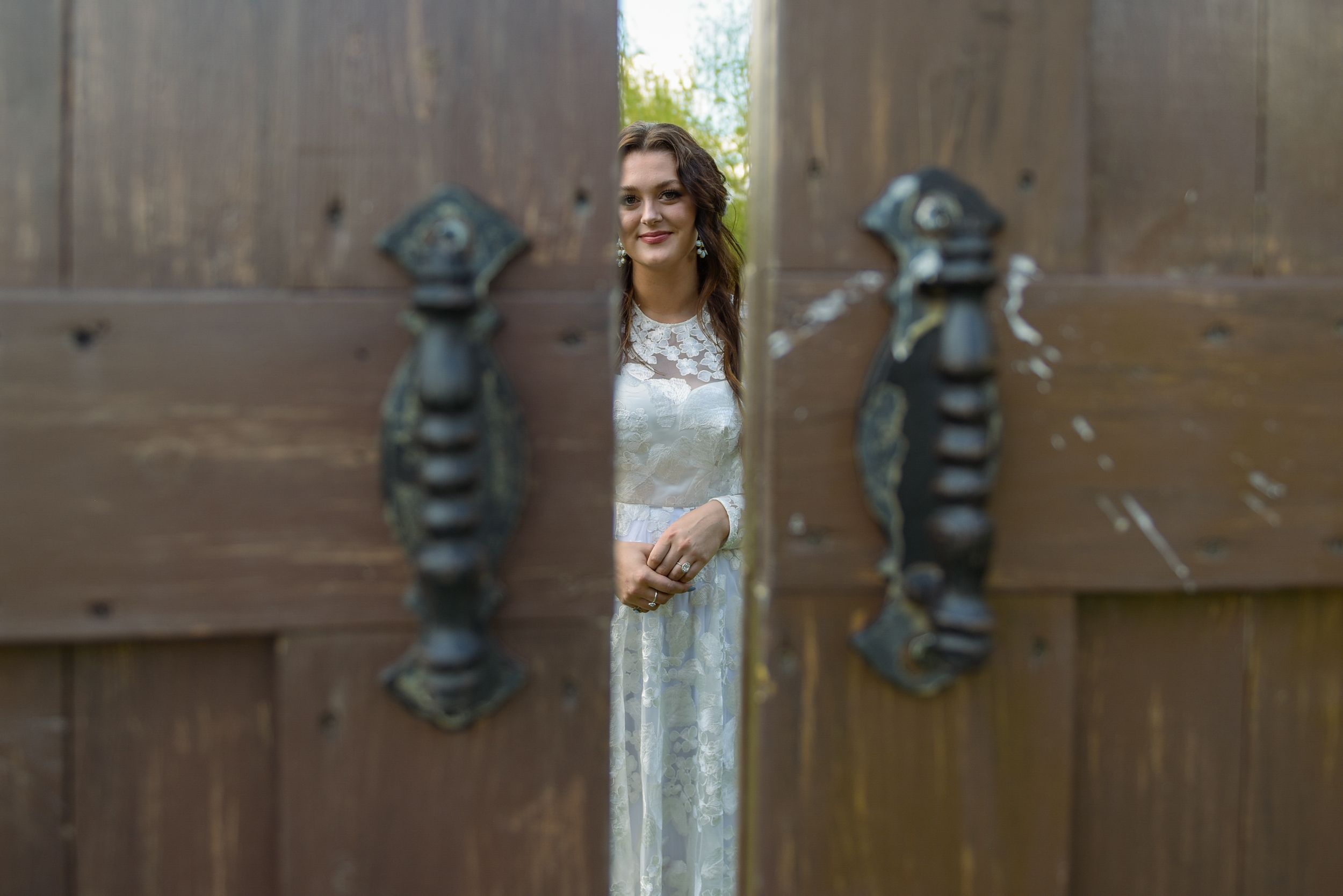 Bride in white lace gown peeks through ornate wooden doors with decorative iron handles.