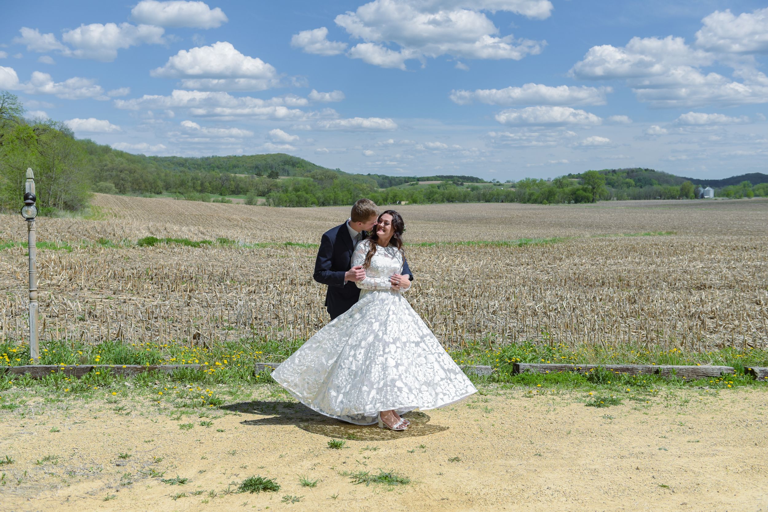 A bride and groom dance outdoors in a rural field landscape under a blue sky with white clouds on their wedding day.