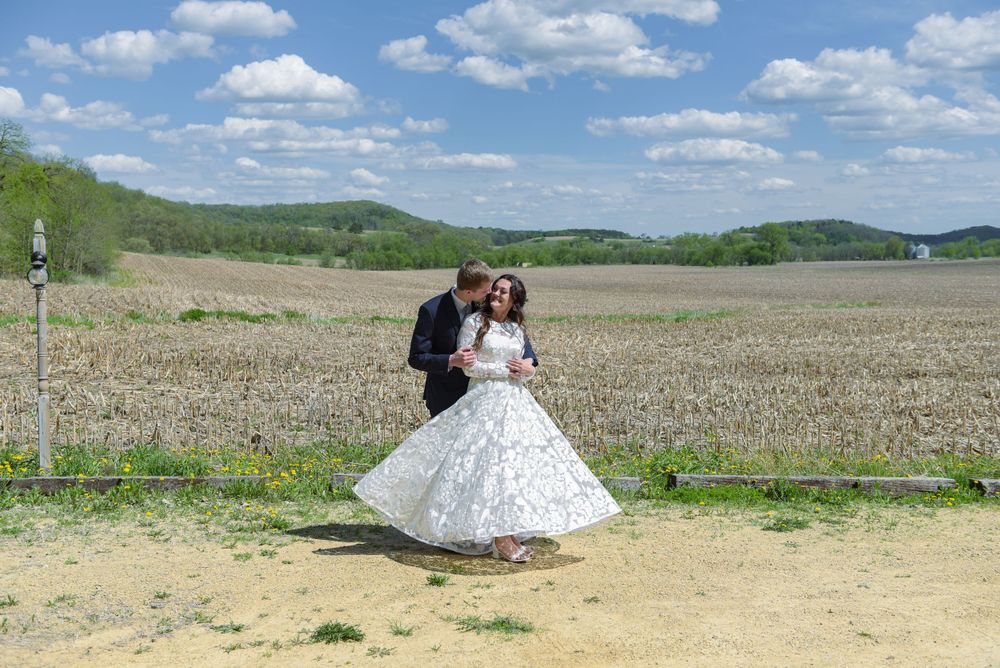 A bride and groom dance outdoors in a rural field landscape under a blue sky with white clouds on their wedding day.
