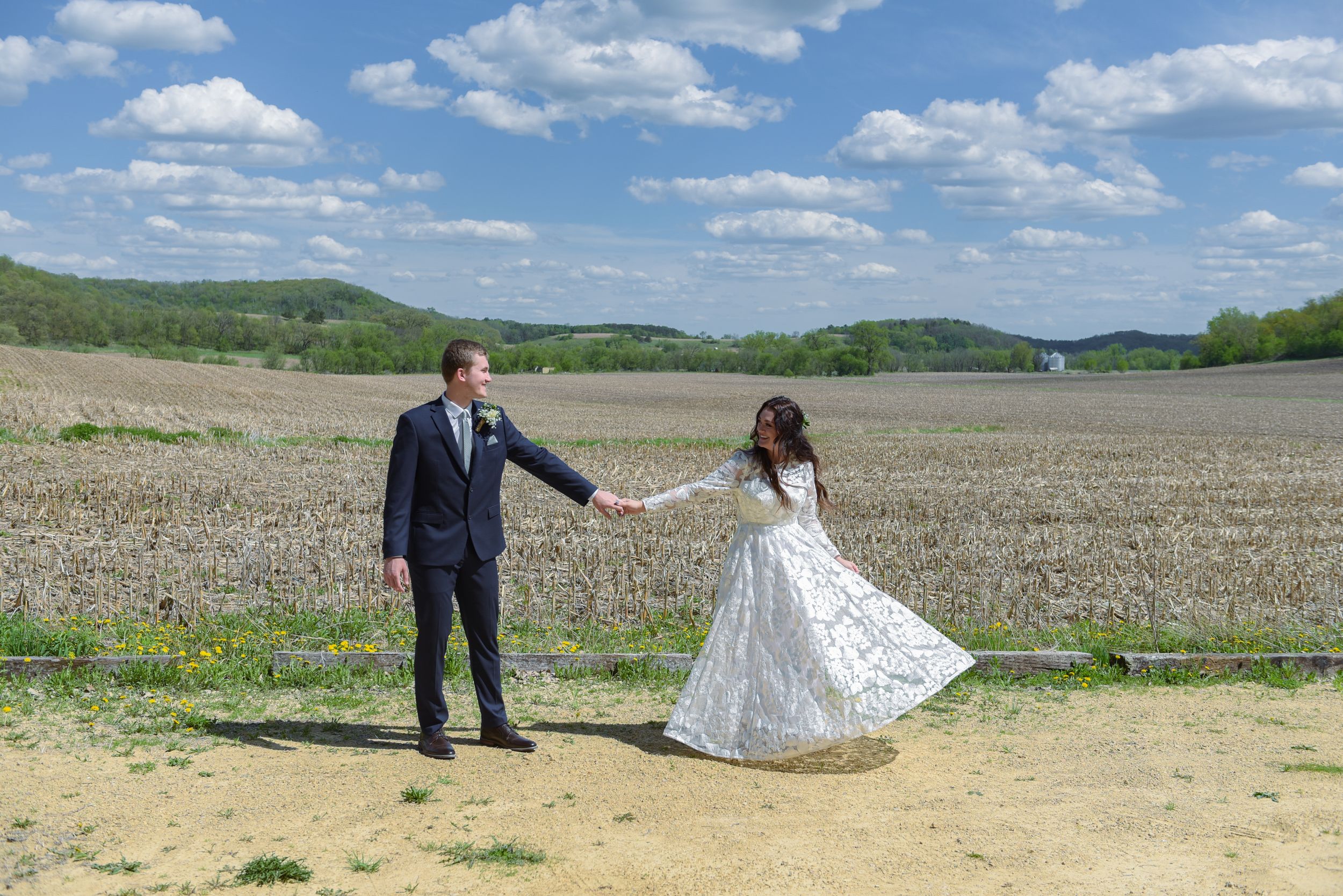 A bride and groom hold hands in a sunny rural field, with rolling hills and blue skies in the background.
