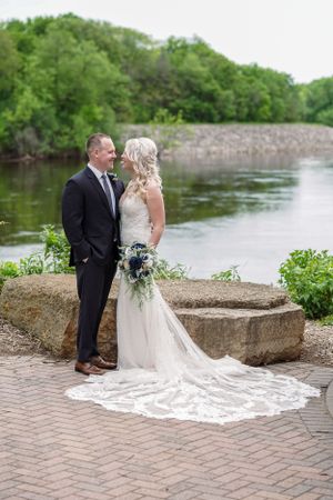 Bride and groom stand facing each other by a river, bride in lace gown with long train holding bouquet, groom in dark suit.