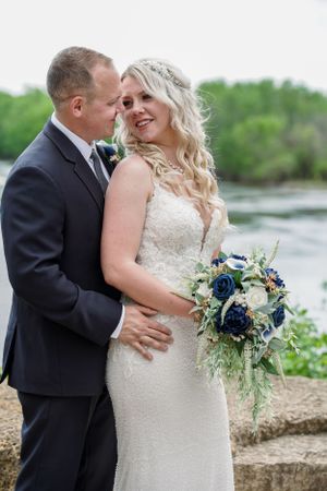Bride in lace gown holds navy bouquet while groom in dark suit embraces her near a scenic waterway.